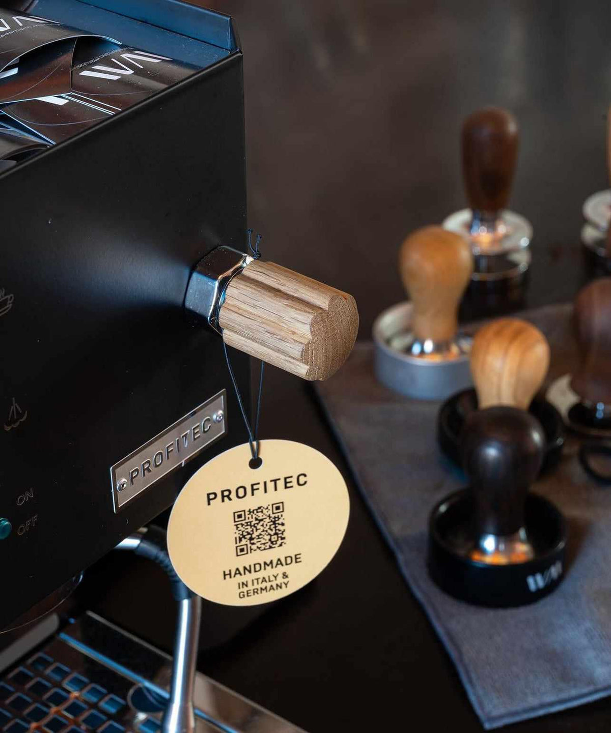 Närbild på en espresso-maskin med trähandtag och en rund etikett med texten "PROFITEC HANDMADE IN ITALY & GERMANY", med flera trä- och metalltamperar i bakgrunden.